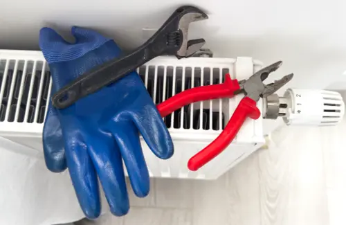 A close-up of a radiator with a blue glove, a black adjustable wrench, and a pair of red-handled pliers sitting on top of a heating system for maintenance.