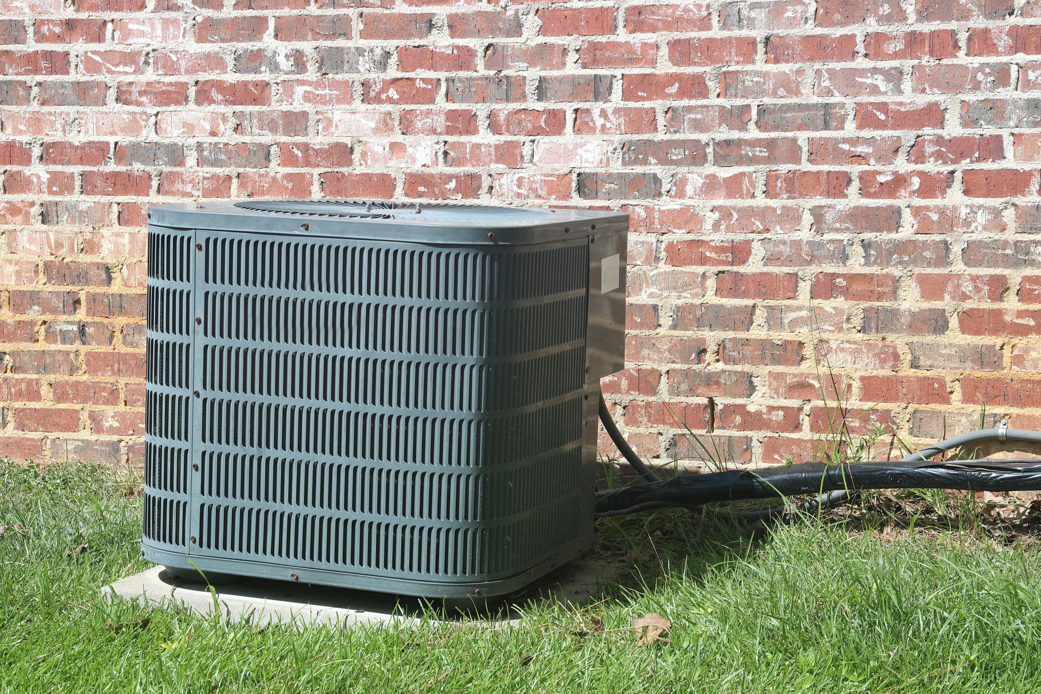 An outdoor air conditioning unit placed on a concrete base, surrounded by grass, and located next to a brick wall.