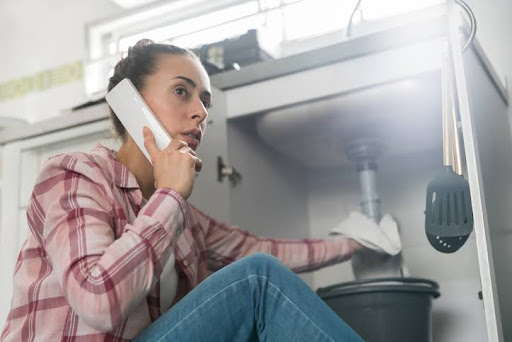 Concerned woman sitting on the kitchen floor makes a phone call while holding a towel under a leaking sink. A bucket is positioned below the exposed plumbing to prevent water damage.